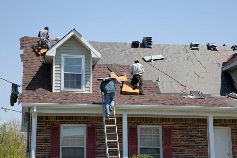 Workers inspect a damaged roof from the ground while a homeowner stands safely near a covered doorway.